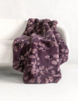 A plush throw blanket with a dusty purple and mauve floral pattern, draped over a white armchair in a bright, minimalist room.