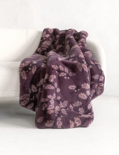 A plush throw blanket with a dusty purple and mauve floral pattern, draped over a white armchair in a bright, minimalist room.