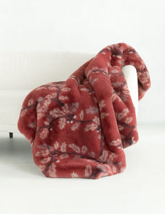 A plush throw blanket draped over a white armchair in a bright, minimalist room. The blanket is a dusty rust red color with a lighter beige leaf pattern.