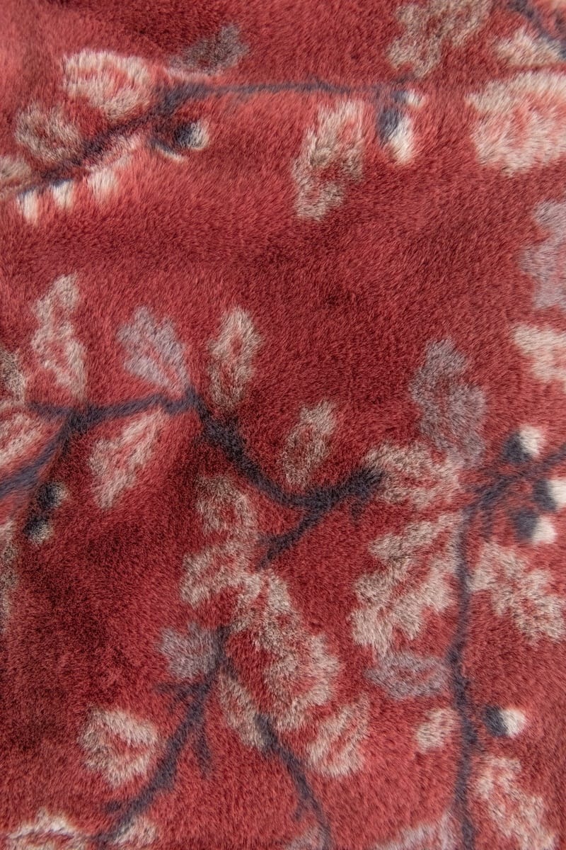 A close-up of a plush, textured fabric with a dusty red base and a light beige and dark floral branch pattern.