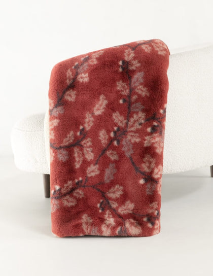 A folded plush throw blanket with a red and beige floral branch pattern, draped over the back of a white armchair against a clean white background.