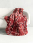A plush throw blanket draped over a white armchair, featuring a dusty red base with a white and dark red floral branch pattern.