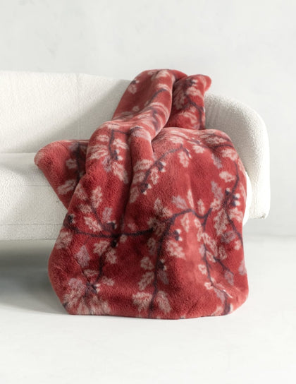 A plush throw blanket draped over a white armchair, featuring a dusty red base with a white and dark red floral branch pattern.