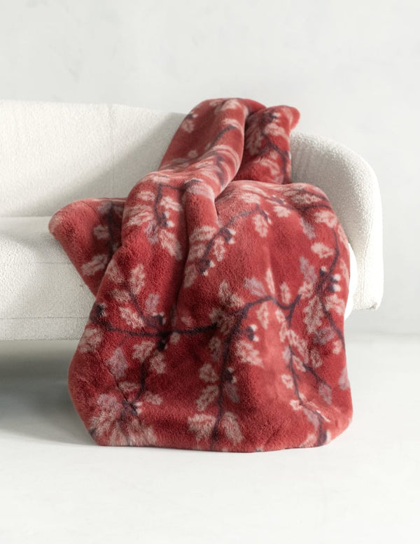 A plush throw blanket draped over a white armchair, featuring a dusty red base with a white and dark red floral branch pattern.