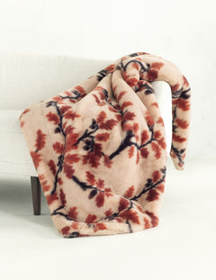 A plush throw blanket draped over a white upholstered chair, featuring a warm beige base with a branch pattern in rust red and dark brown.