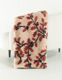 A rectangular throw blanket with a soft plush texture, featuring a pattern of reddish-brown leaves and dark branches on a light beige background, draped over the back of a white textured armchair.