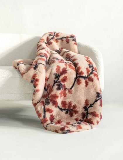 A plush throw blanket with a warm beige base and a rust-red and black leafy branch pattern, draped over a white upholstered armchair in a bright, minimalist room.