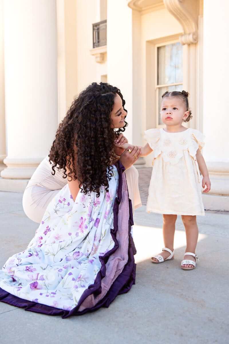 A woman kneels with a Dusty Plum Wildflower Blanket beside a girl in a cream dress, enjoying a sunny day outdoors.