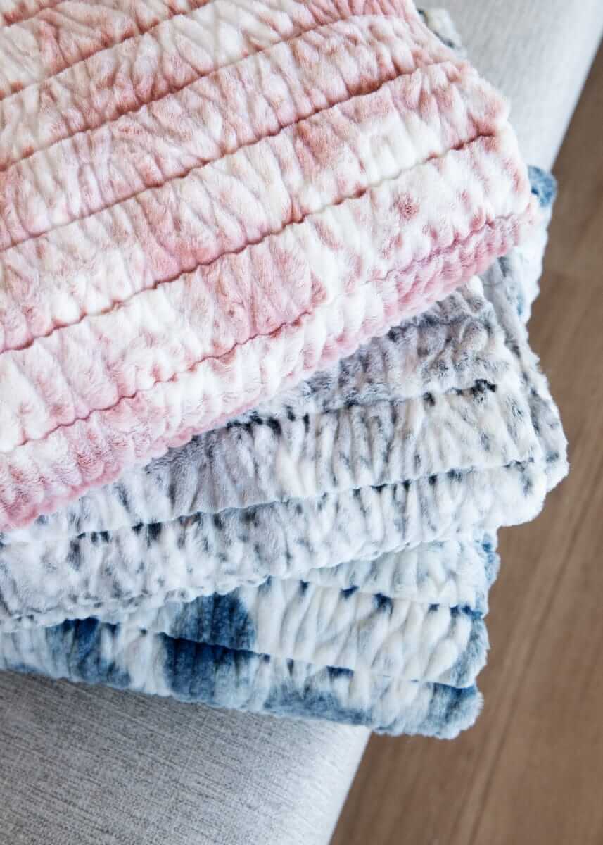 Plush Minky Couture blankets stacked on a sofa, showing layered pink, gray, and blue patterned fabrics.