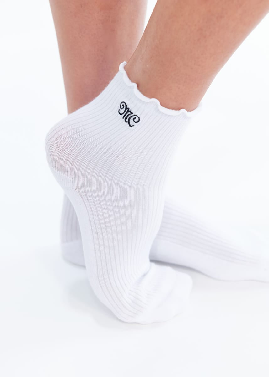 White ankle socks with a scalloped ruffle edge and a black embroidered monogram logo on the cuff, shown on a person's feet against a white background.
