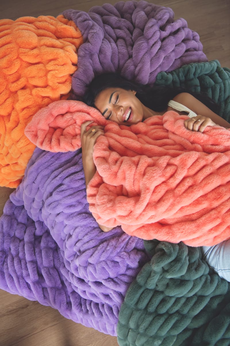 A woman lying on a large chunky knit blanket in a cozy indoor setting. Featuring multiple blankets that are orange, purple, coral pink, and dark green.