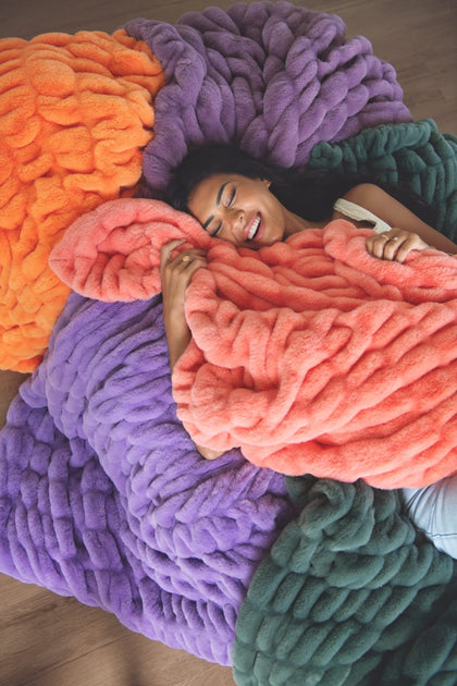A woman lying on a large chunky knit blanket in a cozy indoor setting. Featuring multiple blankets that are orange, purple, coral pink, and dark green.