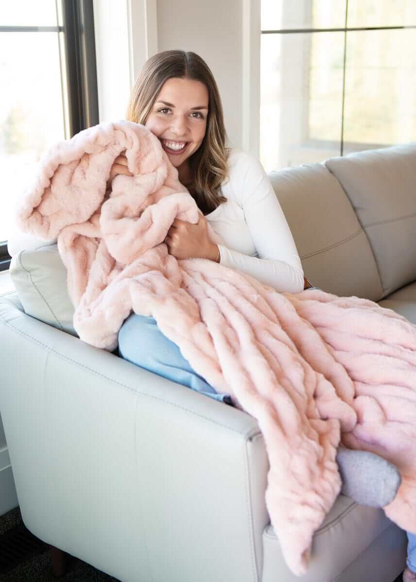 A woman smiling while wrapped in the Hugs Peach Whip Blanket on a sofa, showcasing its soft, cozy texture.