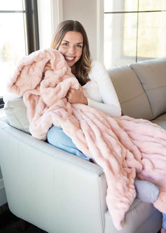 A woman smiling while wrapped in the Hugs Peach Whip Blanket on a sofa, showcasing its soft, cozy texture.