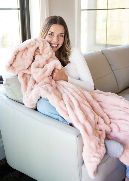 A woman smiling while wrapped in the Hugs Peach Whip Blanket on a sofa, showcasing its soft, cozy texture.