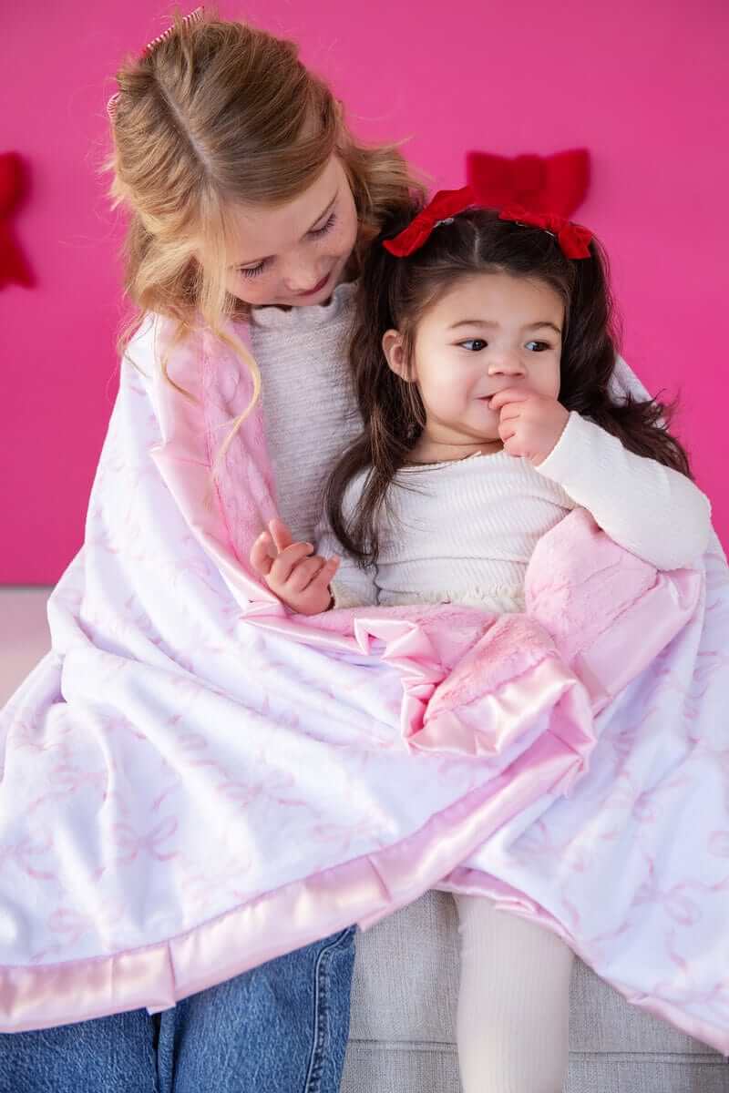 Two children cozy in a blanket featuring Pink Ribbon Bows, set against a vibrant pink background.