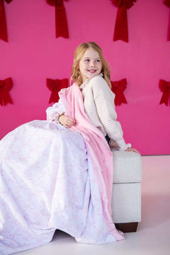 A girl sitting on a couch wrapped in a Pink Ribbon Bows blanket, featuring pink ribbons on a white background.