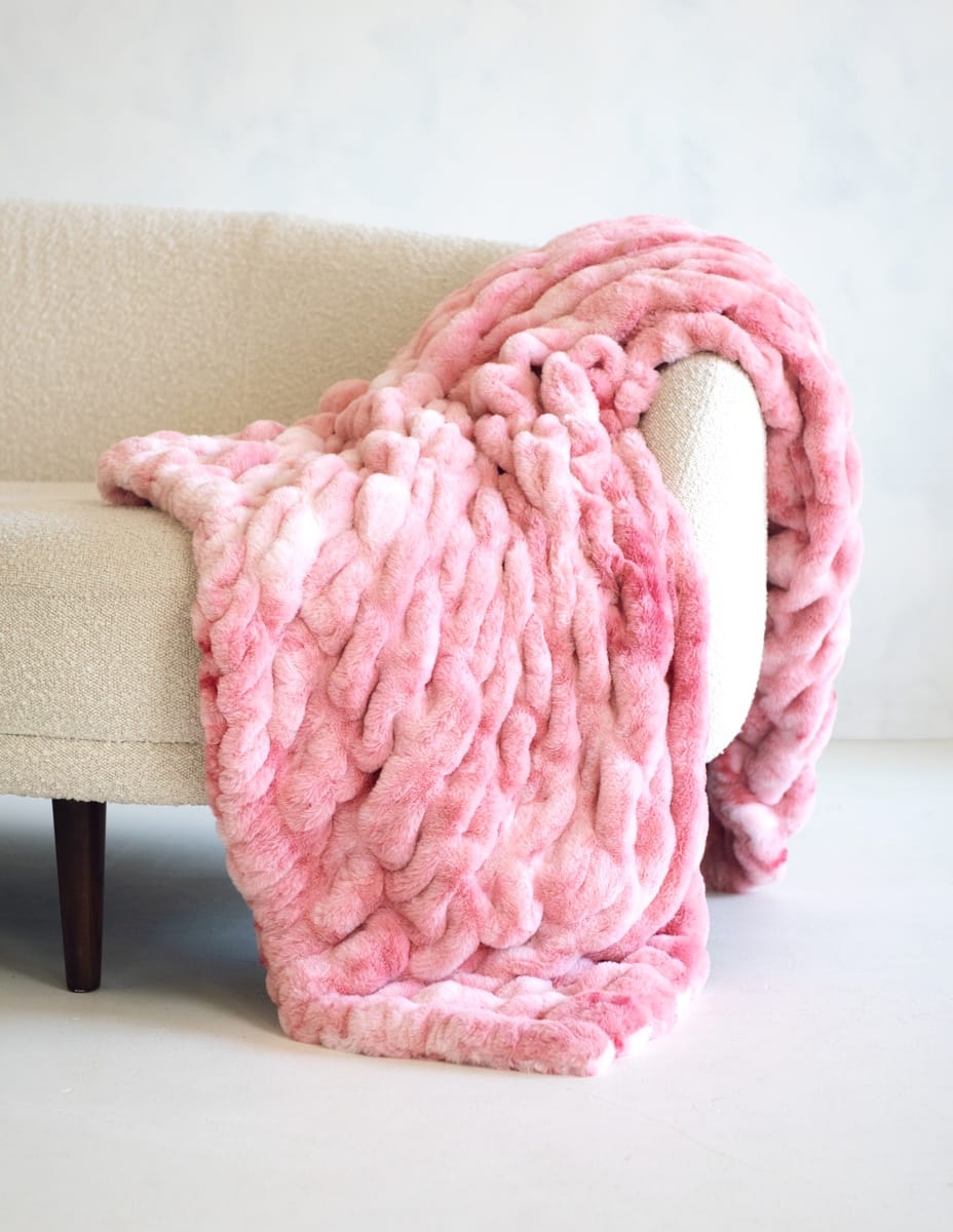 A chunky, Minky Couture Hugs Cotton Candy Pink blanket draped over a light beige upholstered armchair in a bright, minimalist room.