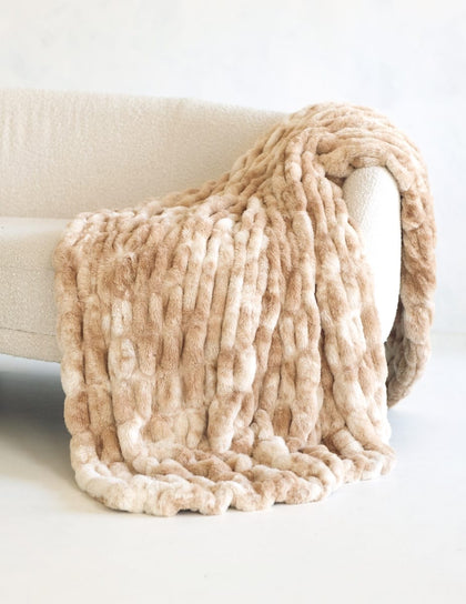 Chunky Hugs Cotton Candy Caramel blanket in a warm beige and cream colorway, draped over the arm of a light-colored sofa.