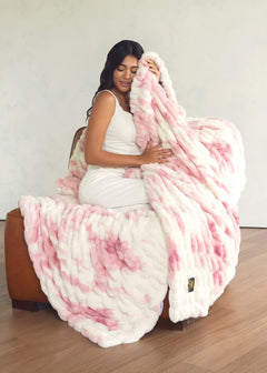 A woman in a white dress sits on a brown leather armchair, wrapped in a Minky Couture throw blanket with a pink and white tie-dye pattern.