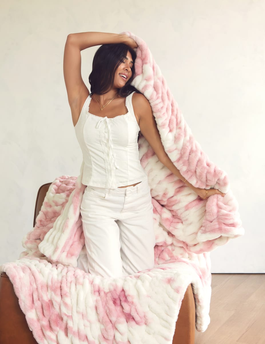 A woman in a white sleeveless top and white pants sits on a brown chair, holding up a soft Minky Couture blanket in pink and white against a plain light wall.