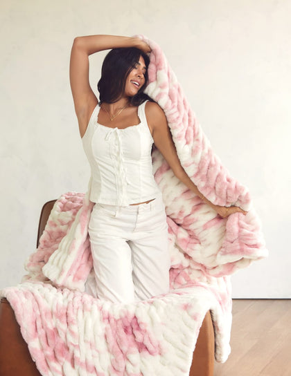 A woman in a white sleeveless top and white pants sits on a brown chair, holding up a soft Minky Couture blanket in pink and white against a plain light wall.