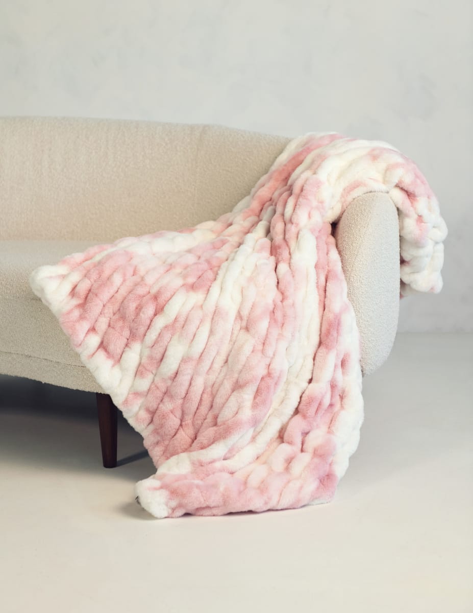 A soft throw blanket in a pink and white marbled pattern, draped over the arm of a light beige upholstered armchair.