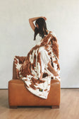 A woman sits on a brown leather armchair draped in a fluffy Minky Couture throw blanket with a brown and white cow-print pattern.