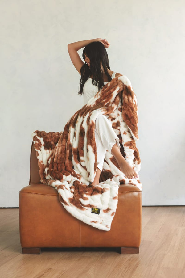A woman sits on a brown leather armchair draped in a fluffy Minky Couture throw blanket with a brown and white cow-print pattern.
