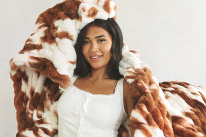  woman with dark hair is wrapped in a plush minky blanket with a brown and white cow print pattern, wearing a white top against a plain light background.