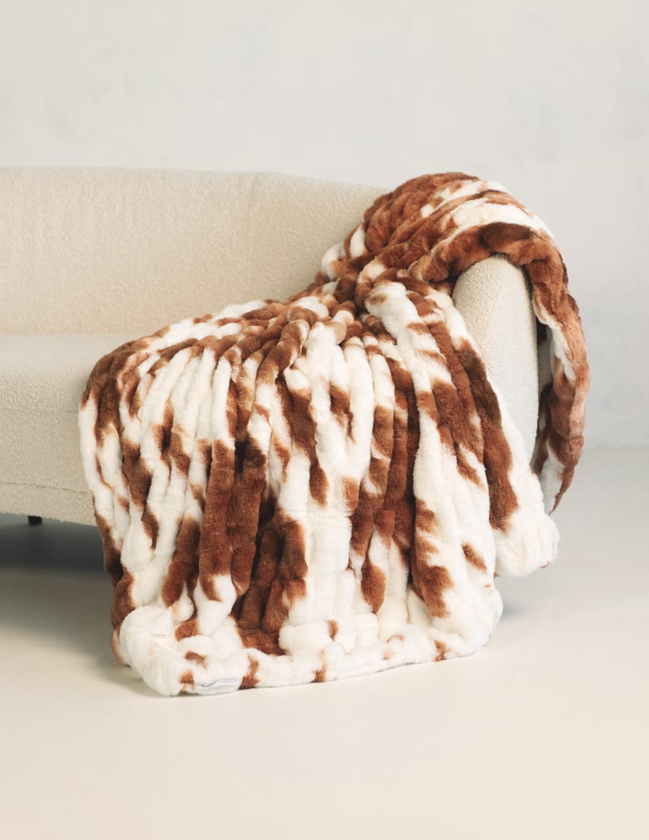 A plush throw blanket with a brown and white cow-like pattern draped over the arm of a light beige sofa in a minimalist indoor setting.
