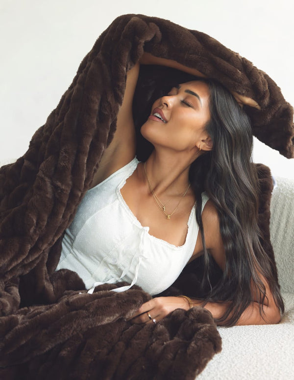 A woman with long dark hair reclines under a plush, thick brown throw blanket, wearing a white camisole top and a delicate gold necklace.