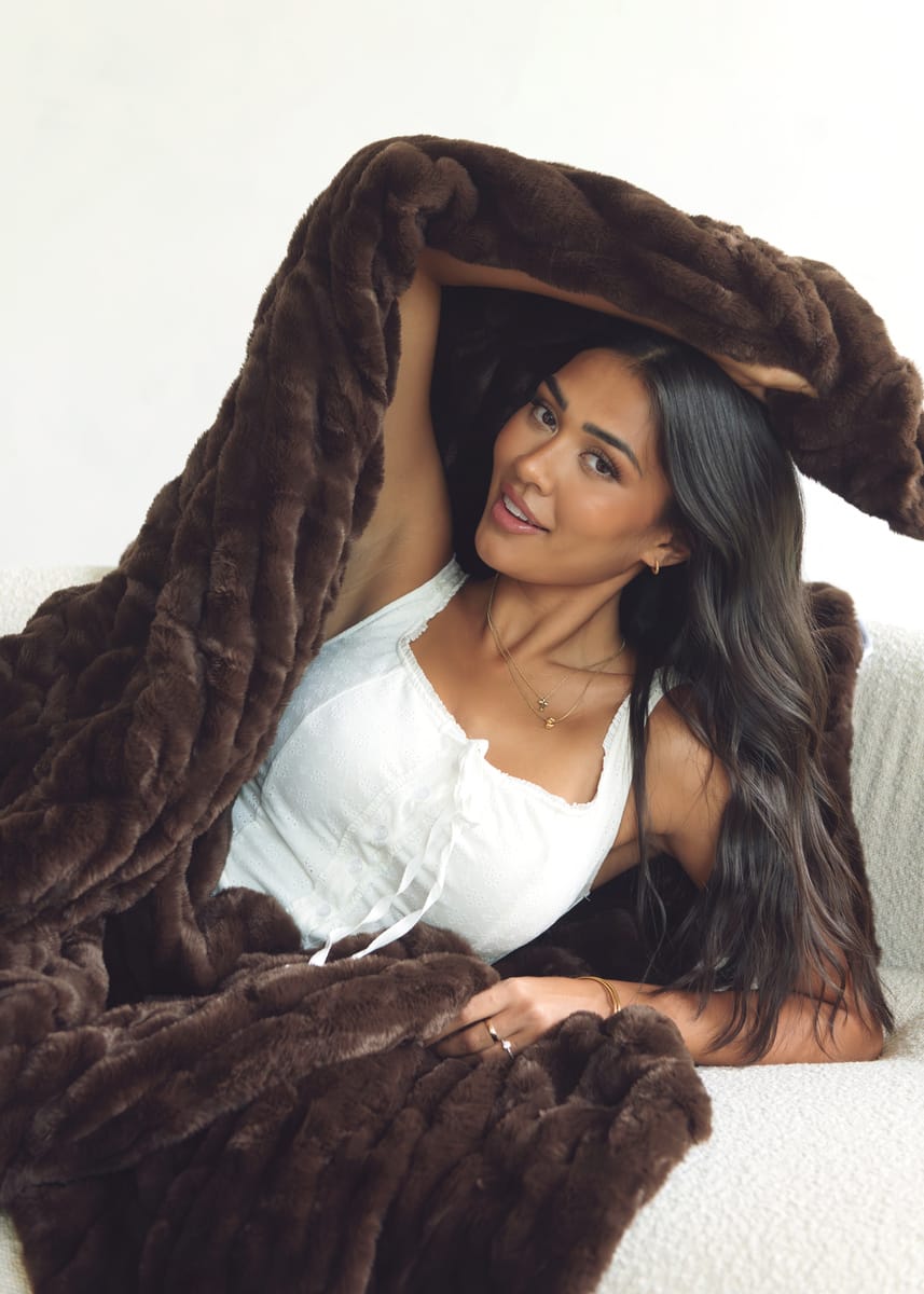 A woman with long dark hair lies on a white textured sofa, wrapped in a plush, chunky dark brown Minky Couture throw blanket. She is wearing a white camisole top and a delicate gold necklace.