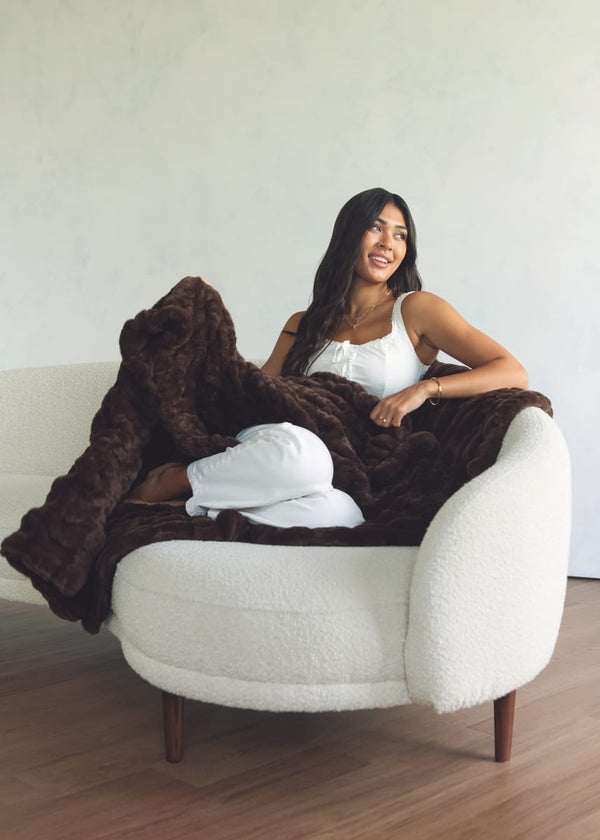 A woman wearing a white camisole and white pants sits on a cream boucle armchair, wrapped in a plush dark brown Minky Couture throw blanket against a light textured wall and wood floor.