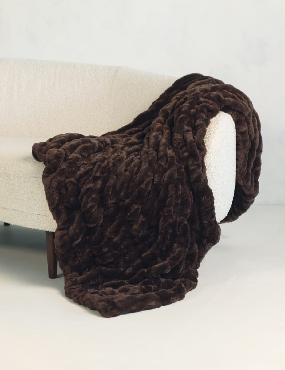 A chunky, plush Minky Couture throw blanket in dark brown draped over a cream-colored armchair against a light gray background.