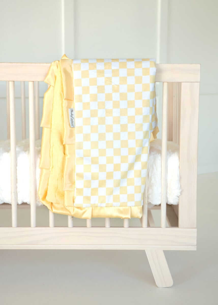 Minky Couture yellow and white Checkered Banana baby blanket draped over a light wood crib.