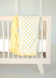 Minky Couture yellow and white Checkered Banana baby blanket draped over a light wood crib.