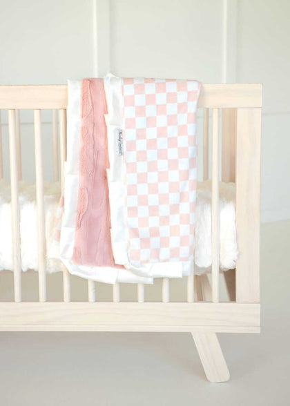 Pink and white Checkered Blush Minky Couture baby blanket draped over a light wood crib railing.