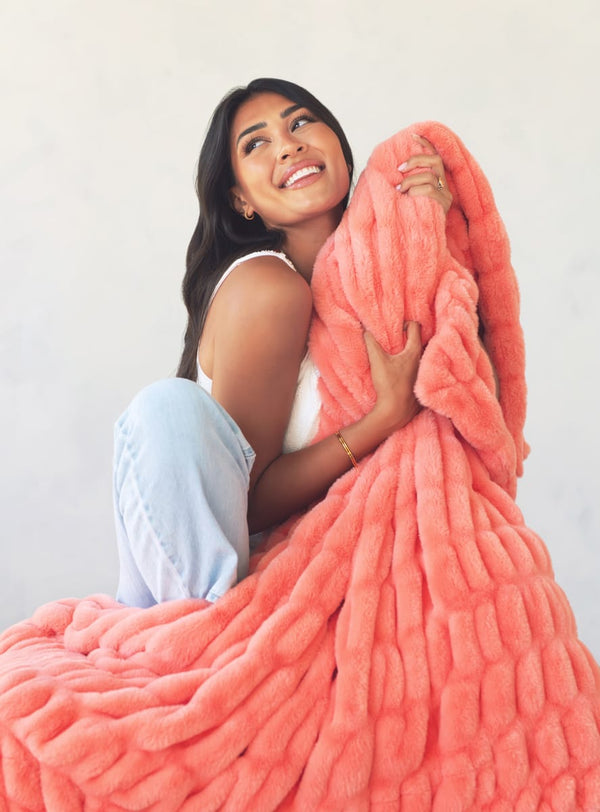 A woman smiling and hugging a large, chunky coral-pink plush blanket against a plain light background.