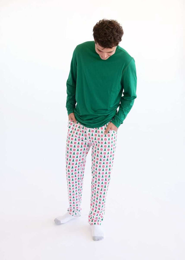 Man wearing a green long-sleeve top and white Christmas tree pajama pants, standing against a white background.