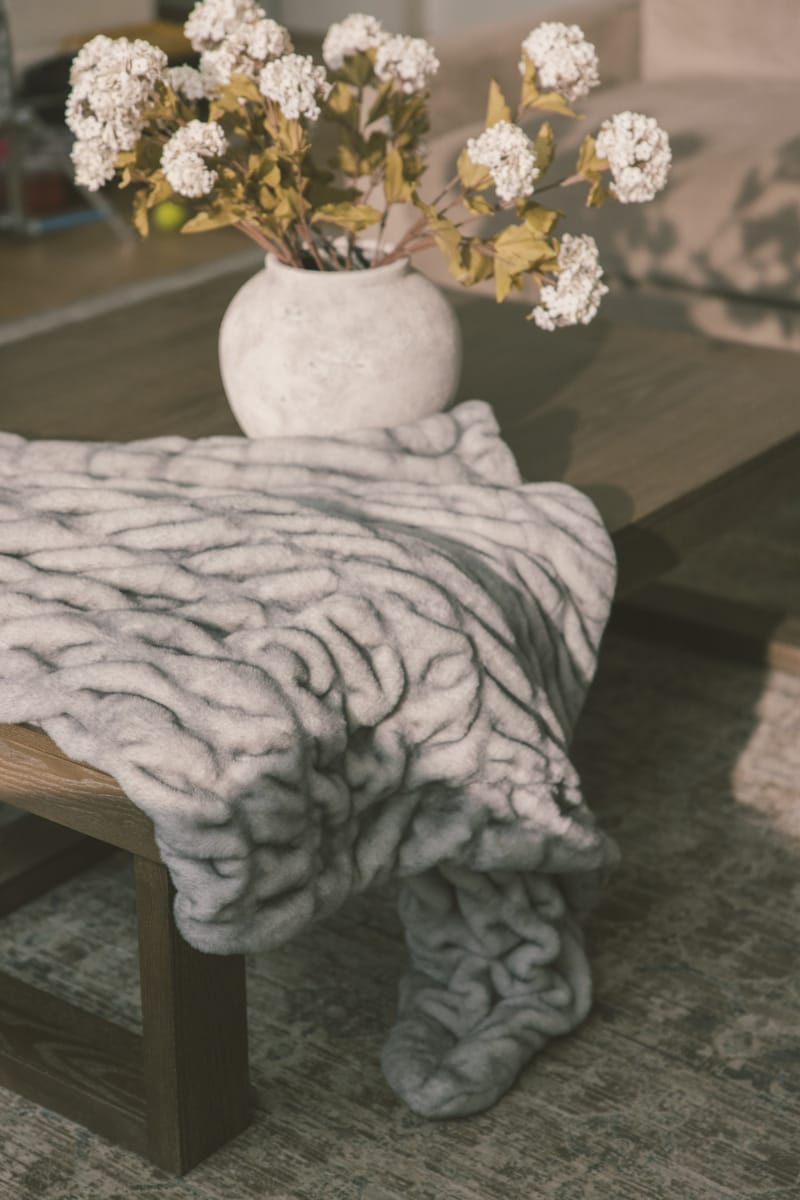 Gray textured blanket draped over a wooden chair with a vase of white flowers on a table.