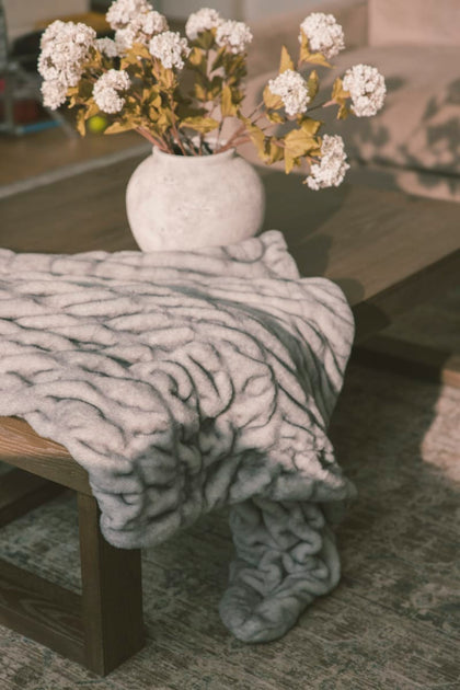 Gray textured blanket draped over a wooden chair with a vase of white flowers on a table.