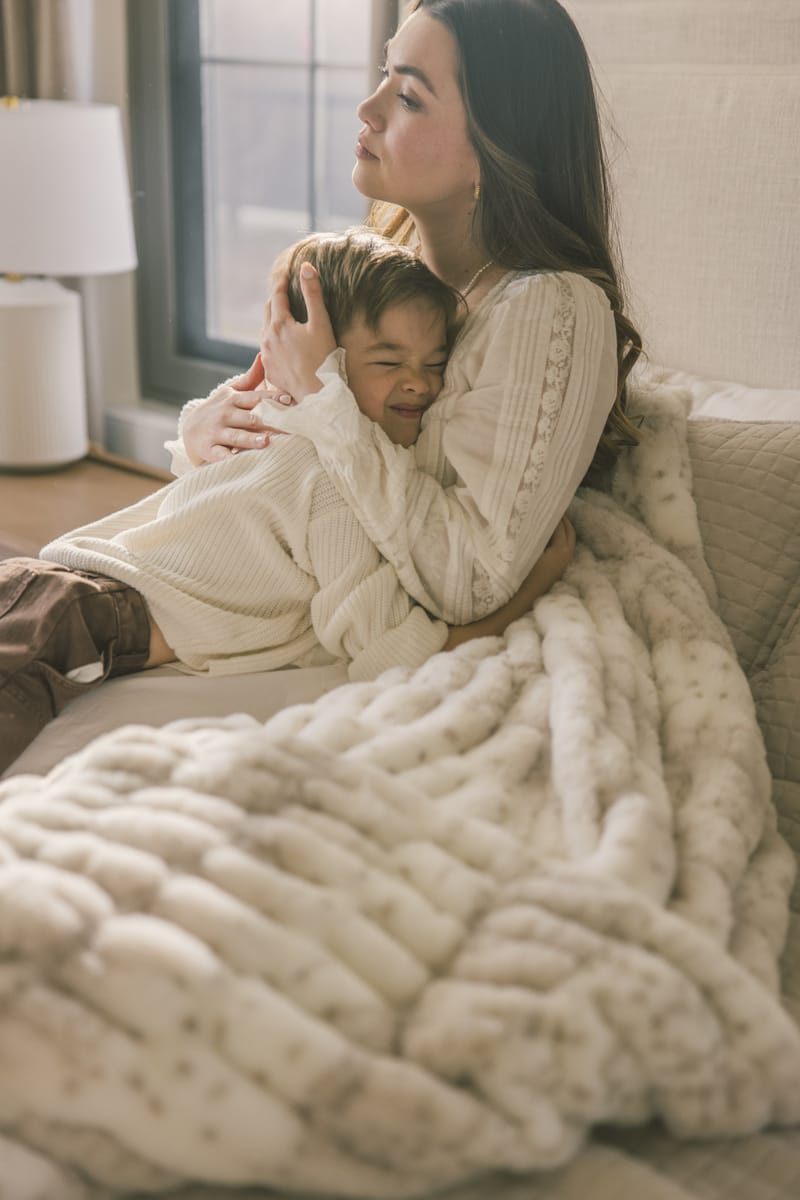 Woman and child sitting on a couch under a large white blanket in a cozy living room.