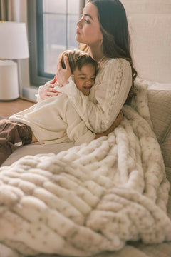Woman and child sitting on a couch under a large white blanket in a cozy living room.