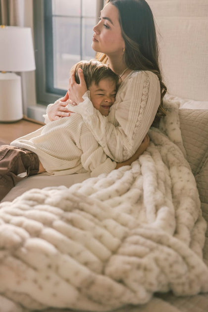 Woman and child sitting on a couch under a large white blanket in a cozy living room.