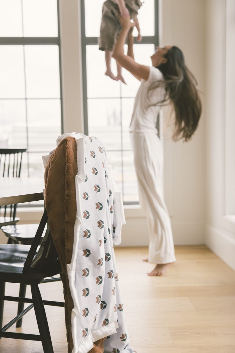 Woman lifting a child in a bright room with large windows and a chair with a patterned blanket draped over it.