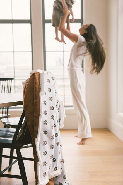 Woman lifting a child in a bright room with large windows and a chair with a patterned blanket draped over it.