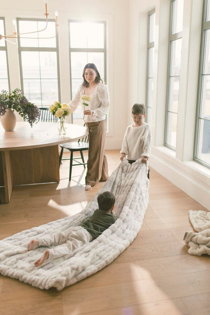 Two little boys playing with a large cream blanket in the kitchen.