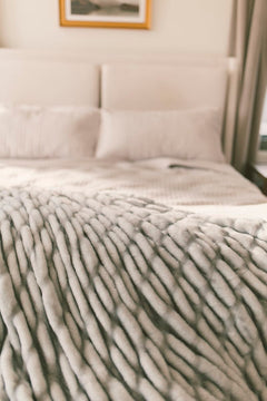 Close-up of a textured gray blanket on a bed with pillows in a softly lit bedroom.
