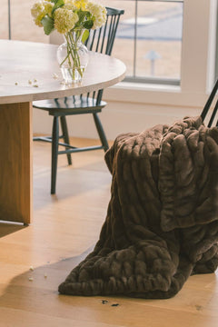 Hugs Hot Fudge Blanket draped over a chair in a stylish indoor setting, showcasing its cozy brown design.